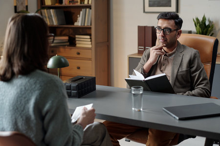 Woman sitting across from man holding notebook and pen, listening attentively during therapy session in professional office settingの写真素材
