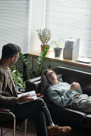 Woman lying on couch talking to man holding notebook during therapy session in office with plants and window blinds in backgroundの写真素材
