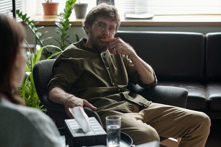 Man sitting in therapy session holding tissue box and looking thoughtfully at therapist, indoor setting with plants and sofa in backgroundの写真素材