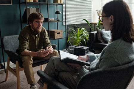 Man sitting in therapy session talking to woman therapist holding notebook and pen, both seated in modern office setting with plants visibleの写真素材