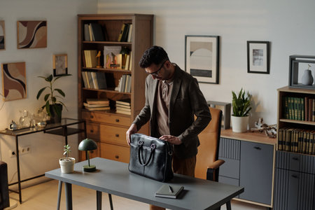 Man standing in office opening black briefcase on desk preparing for therapy session with bookshelves and framed artwork visible in backgroundの写真素材
