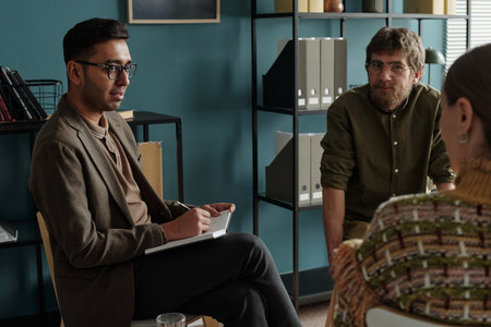 Man holding notebook leading therapy session with man and woman listening attentively in office settingの写真素材