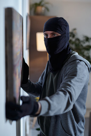 Burglar wearing balaclava and gloves removing painting from wall, looking focused while committing theft in residential home, partial side viewの写真素材