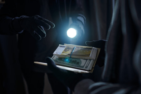 Two adult men wearing gloves exchanging stack of euro banknotes in dark room, one holding flashlight...