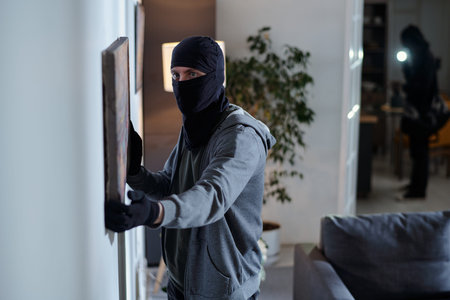 Man wearing black balaclava and gloves stealing painting from wall while another masked searching...