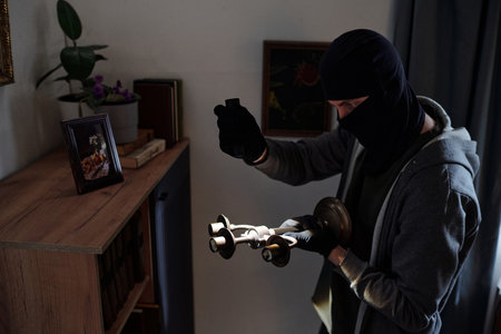 Male wearing balaclava and gloves holding candlestick and flashlight standing near bookshelf appearing to commit burglary in residential houseの写真素材