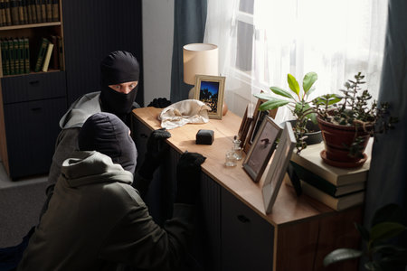 Two men wearing black ski masks crouching near cabinet rummaging through drawers in daylight living...