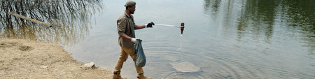 Young adult Black man cleaning riverbank, holding trash bag in one hand and using metal grabber to...