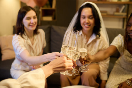 Group of young multiethnic women sitting together clinking champagne glasses celebrating bachelorette and upcoming marriage, smiling and enjoying festive moment in cozy indoor settingの写真素材