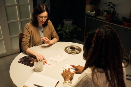 Caucasian young woman choosing card at tarot reading with Black female mystic, sitting at round table, both focusing on cards, notebook and glass of water on tableの写真素材