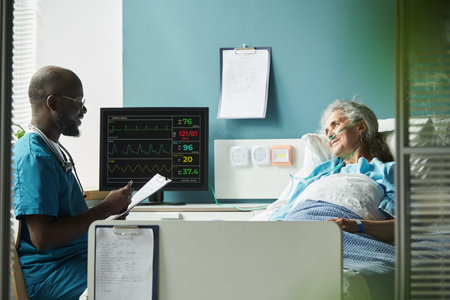 Senior Caucasian woman lying in hospital bed connected to medical monitor, Black middle aged male doctor sitting nearby holding clipboard and engaging in conversation with patientの写真素材