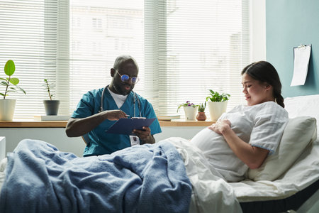 Black middle aged male doctor writing on clipboard while sitting beside pregnant Caucasian young woman lying in hospital bed, both interacting during prenatal checkupの写真素材