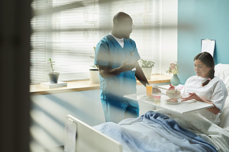 Pregnant Caucasian woman sitting in hospital bed eating meal while Black middle aged male nurse standing beside bed talking to her, medical tray with food and drink on her lapの写真素材
