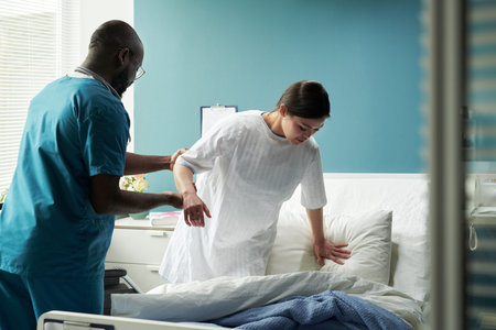 Middle aged Black male nurse assisting young Caucasian woman getting out of hospital bed in maternity clinic, healthcare professional supporting patient during recovery processの写真素材