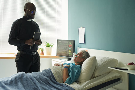 Black middle aged man holding rosary and book standing beside hospital bed, visiting senior...