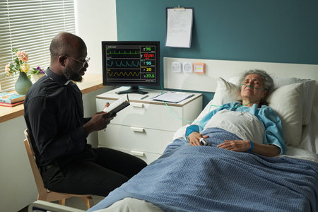 African American middle aged male priest sitting beside hospice bed reading prayers from Holy Bible to senior Caucasian woman resting with eyes closed, while providing end of life careの写真素材