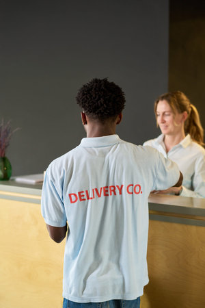 Black young man standing at reception desk interacting with young woman receptionist, delivery...