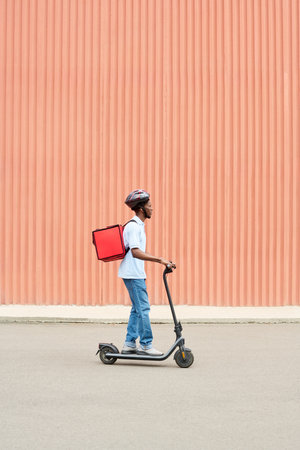 Black young man riding electric scooter carrying insulated delivery backpack wearing helmet moving along urban street in front of large corrugated orange wallの写真素材