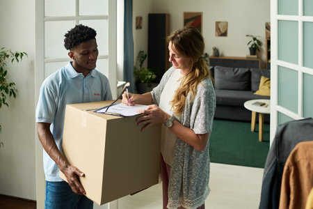 Woman signing delivery form while Black young man holding large cardboard box, both standing in...