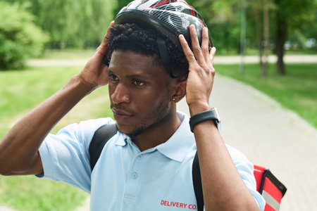Young Black man adjusting bicycle helmet outdoors, wearing delivery backpack and uniform shirt,...