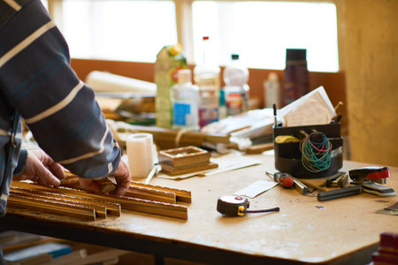 Man working with wooden slats on cluttered workshop table, hands arranging materials near measuring...