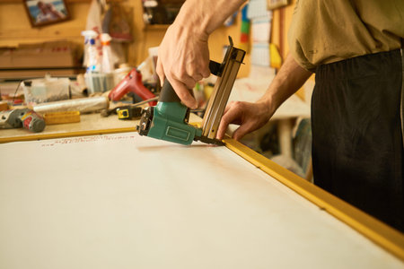 Man using electric staple gun to fasten canvas onto wooden frame in workshop, hands gripping tool...
