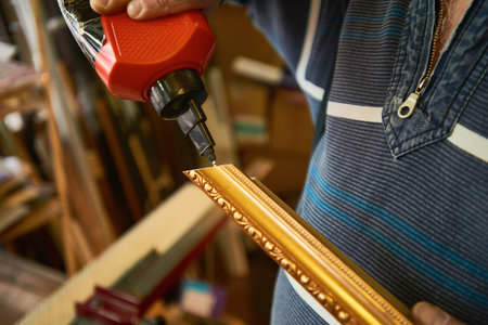 Man assembling ornate picture frame using electric screwdriver, focusing on precise alignment of decorative molding in woodworking or framing workshopの写真素材