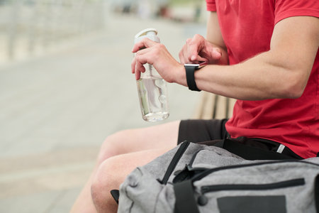 Young man sitting outdoors checking smartwatch while holding water bottle gym bag resting on lap forearms and hands visible preparing for workoutの写真素材