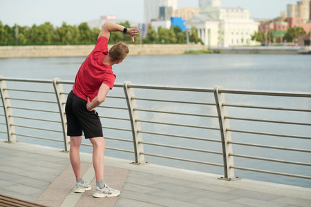 Young man stretching side body outdoors near river standing on paved walkway with hand on hip...