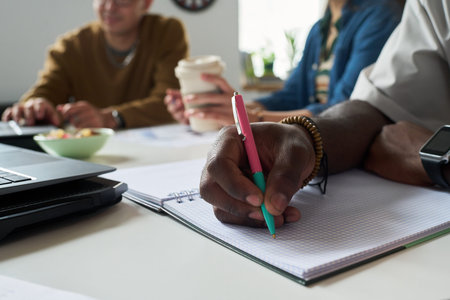 Black man writing in notebook with pen while sitting at table with multiethnic group of adults in...