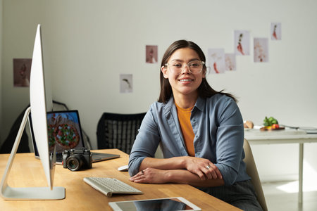 Portrait of woman sitting at desk smiling at camera, using desktop computer with digital tablet and...
