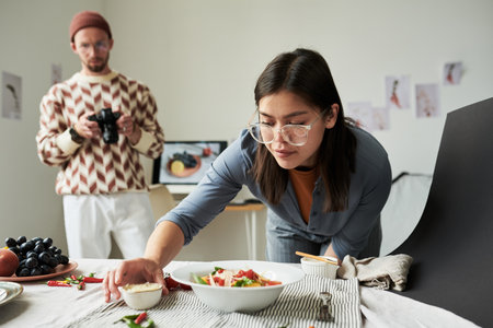 Asian woman arranging food on table while man with beard photographing scene in background, creative workspace with food styling setup visibleの写真素材