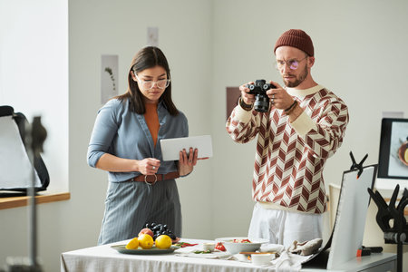 Man photographing food on table with digital camera while woman holding tablet standing beside him,...
