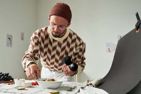 Man wearing glasses holding camera styling food on table preparing for food photography session in...