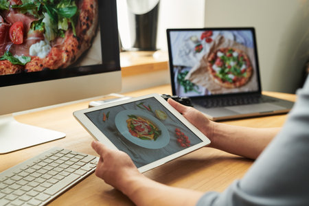 Woman holding tablet displaying food photo while sitting at desk with computer and laptop showing similar images, working on digital content creationの写真素材