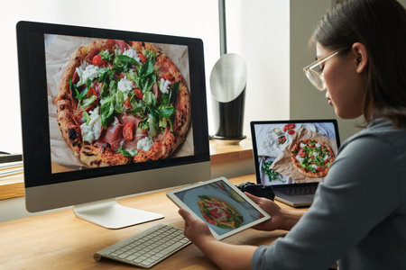 Asian woman sitting at desk using digital tablet while looking at large computer monitor displaying...