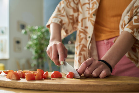 Young girl slicing cherry tomatoes on wooden cutting board in kitchen, hands holding knife and preparing fresh vegetables for cooking or saladの写真素材