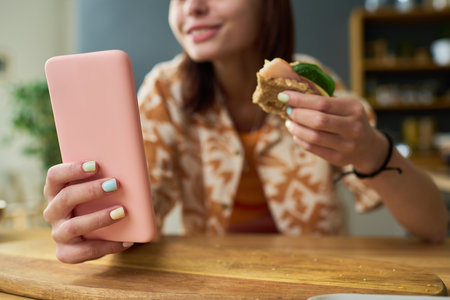Young girl holding smartphone with pastel painted nails, smiling while eating sandwich in kitchen, focusing on device screen and enjoying meal during breakの写真素材