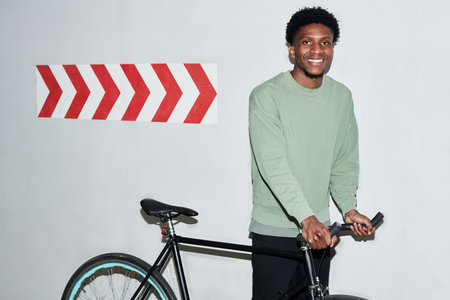 Young man smiling while standing with bicycle near wall with red and arrow sign, looking slightly...