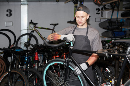 Young man adjusting handlebars on bicycle in workshop, wearing gloves and apron, surrounded by...