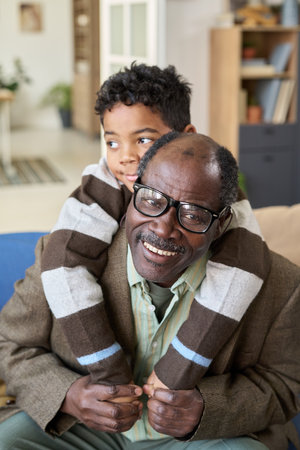 Black senior man sitting on sofa smiling while boy hugging him from behind, both looking happy and relaxed in home setting, showing multigenerational family bondingの写真素材