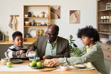 Senior Black man sitting at kitchen table with two Black boys, pouring milk into bowl while boys preparing breakfast, family members interacting during morning meal at homeの写真素材