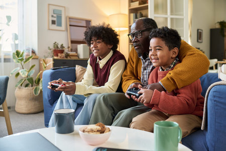 Black senior man sitting on sofa playing video game with two Black boys, smiling and holding...