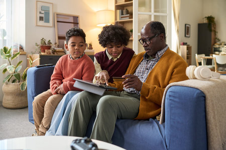 Senior man sitting on sofa with two children, holding credit card and showing tablet, children...