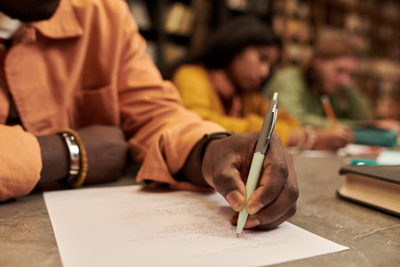 Young man writing on paper at desk in library with multiethnic young people studying in background, focusing on hand holding pen and academic environmentの写真素材