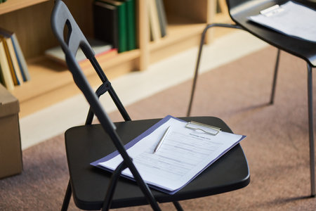 Empty black chair holding clipboard with group therapy paperwork and pen resting on seat, background showing additional chairs and bookshelf, no people in sceneの写真素材