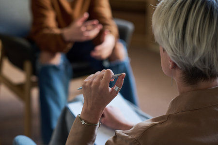 Adult woman holding pen and taking notes while listening to young man gesturing during group therapy session, both seated and engaged in conversationの写真素材