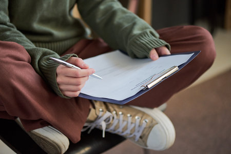 Young woman sitting cross legged holding clipboard and pen writing notes during group therapy session, only hands and lower body , focus on activityの写真素材