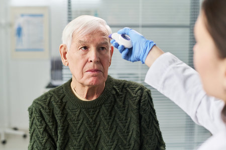 Senior man sitting while female doctor measuring forehead temperature using digital infrared...