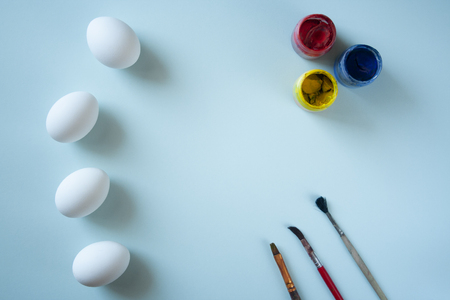 A set of white clean eggs, blue, yellow and red paints and brushes on bright background top view, flat lay. Easter symbols. Easter eggs, painting eggs, easter illustration image.  Easter entertainment concept, easter tradition. Copyspace, place for textの写真素材